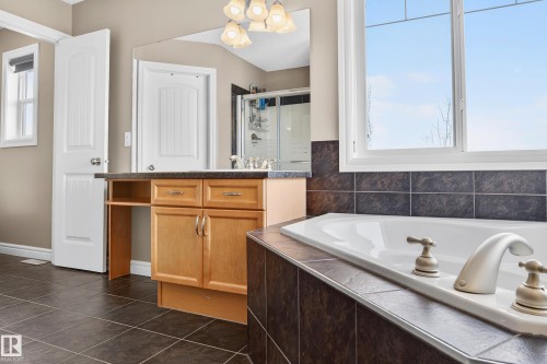 Bathroom featuring a corner soaking tub with deck-mounted fixtures, wood-finish vanity with built-in storage and countertop, tile flooring, and a large window - 8477 Sloane Crescent, Edmonton, AB - Indoor Photo Showing Bathroom