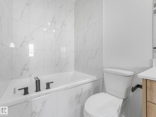 Built-in bathtub with matte black fixtures, surrounded by large format white tile with grey veining, featuring a white toilet and a partial wood-finish vanity with a light-colored countertop - 272 Munn Way, Leduc, AB - Indoor Photo Showing Bathroom