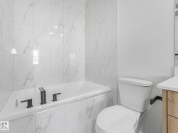 Built-in bathtub with matte black fixtures, surrounded by large format white tile with grey veining, featuring a white toilet and a partial wood-finish vanity with a light-colored countertop - 