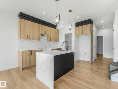 Modern kitchen featuring a central island with a waterfall countertop, light wood-finish cabinetry, white countertops with veining, a black matte faucet, and recessed lighting - 272 Munn Way, Leduc, AB - Indoor Photo Showing Kitchen With Upgraded Kitchen
