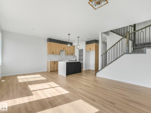 Open-concept living space featuring wood-finish flooring, a modern kitchen with light wood cabinetry and a dark island, marble-patterned backsplash, and a staircase with wrought iron balusters - 272 Munn Way, Leduc, AB - Indoor Photo Showing Kitchen