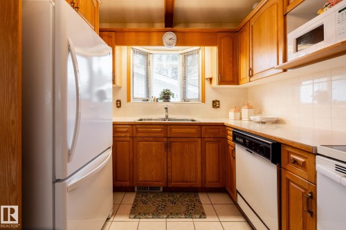 Kitchen featuring wood cabinetry, white tiled flooring, a bay window with blinds, a double basin sink, and white appliances - 8612 68A Street, Edmonton, AB - Indoor Photo Showing Kitchen With Double Sink