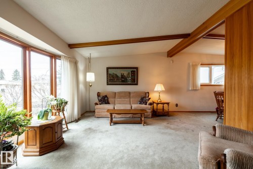 Living area featuring a vaulted ceiling with exposed wood beams, a broad picture window with wood framing, and light-colored wall-to-wall carpeting - 8612 68A Street, Edmonton, AB - Indoor
