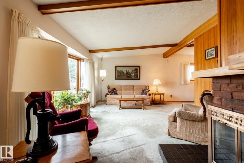Spacious living area featuring a brick fireplace with a metal insert, wood-paneled walls, exposed wood ceiling beams, and light-toned carpeting - 8612 68A Street, Edmonton, AB - Indoor Photo Showing Living Room