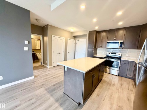 Open-concept kitchen featuring an expansive central island with a light-toned countertop, wood-finish flooring, dark cabinetry, stainless steel appliances, and recessed lighting - 437 5504 Schonsee Drive, Edmonton, AB - Indoor Photo Showing Kitchen