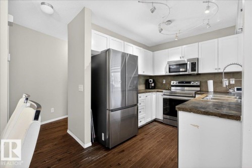 Kitchen featuring dark countertops, stainless steel appliances, white cabinetry, tasteful backsplash, and dark wood finished floors - 47 230 Edwards Drive, Edmonton, AB - Indoor Photo Showing Kitchen With Double Sink