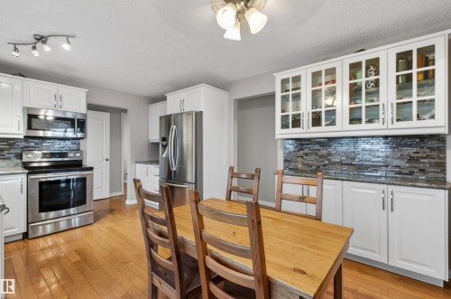 Open-concept kitchen and dining area featuring stainless steel appliances, white cabinetry, wood-finish flooring, a mosaic tile backsplash, and built-in glass-front display cabinets - 17120 103 Street, Edmonton, AB - Indoor Photo Showing Other Room