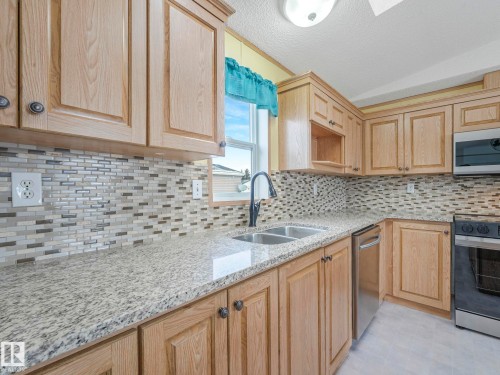 Kitchen featuring granite countertops, a double basin stainless steel sink with a matte black faucet, wood-finish cabinetry, a tile backsplash, and stainless steel appliances - 151 3400 48 Street, Stony Plain, AB - Indoor Photo Showing Kitchen With Double Sink With Upgraded Kitchen