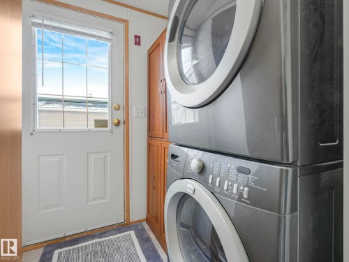 Dedicated laundry area featuring a stacked washer and dryer, wood-finish cabinetry, and a white paneled door with an integrated window - 151 3400 48 Street, Stony Plain, AB - Indoor Photo Showing Laundry Room