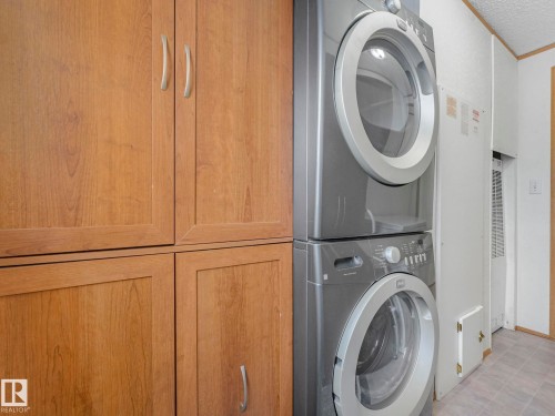 Laundry area featuring wood-finish cabinetry with silver-tone hardware, a stacked washer and dryer, and vinyl flooring - 151 3400 48 Street, Stony Plain, AB - Indoor Photo Showing Laundry Room