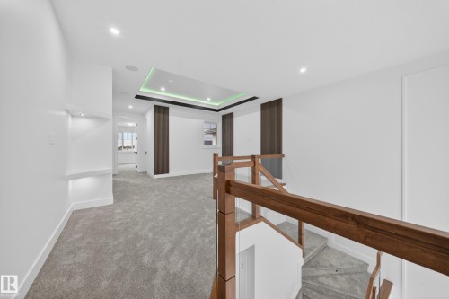 Open landing area featuring gray carpeting, recessed ceiling lighting, a contemporary stair rail with wood-finish top rail and glass panels, a ceiling tray with perimeter lighting, and architectural wall accents - 4005 39 Street, Beaumont, AB - Indoor Photo Showing Other Room