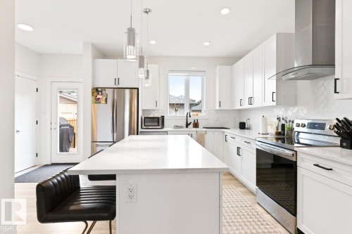 Modern kitchen featuring a large central island, white cabinetry with black hardware, stainless steel appliances, a herringbone tile backsplash, and light wood-finish flooring - 6023 180 Avenue, Edmonton, AB - Indoor Photo Showing Kitchen With Stainless Steel Kitchen With Upgraded Kitchen