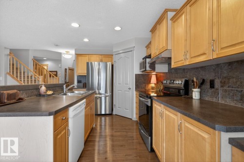 Well-appointed kitchen featuring wood-finish cabinetry, stainless steel appliances, dark countertops, a dark tile backsplash, and recessed lighting - 2178 Haddow Drive, Edmonton, AB - Indoor Photo Showing Kitchen With Double Sink