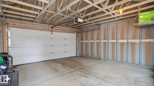Spacious garage featuring a sectional overhead door with an automatic opener, exposed wood ceiling joists, concrete flooring, and a wall-mounted wire storage shelf - 8210 180 Avenue Nw, Edmonton, AB - Indoor Photo Showing Garage