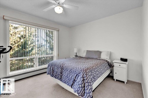 Bright bedroom featuring a large window with vertical blinds, a ceiling fan with integrated lighting, and neutral carpeting - 415 15503 106 Street, Edmonton, AB - Indoor Photo Showing Bedroom