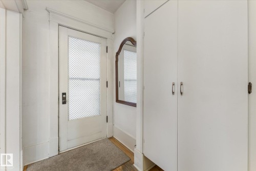 Entryway featuring a white door with a textured glass panel, wood-finish flooring, a built-in storage cabinet with dual doors, and a decorative wall mirror - 10921 126 Street, Edmonton, AB - Indoor Photo Showing Other Room