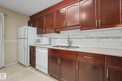 Kitchen featuring wood-finish cabinetry with silver-tone hardware, white subway tile backsplash with accent band, speckled countertops, and a double basin sink with a gooseneck faucet - 1542 54 Street, Edmonton, AB - Indoor Photo Showing Kitchen With Double Sink
