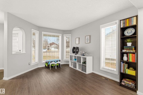 Spacious room featuring wood-finish flooring, a prominent bay window with white blinds, and a built-in dark wood shelving unit - 14755 131 Street Nw, Edmonton, AB - Indoor