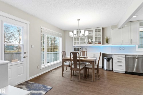 Open-concept kitchen and dining area featuring wood-finish flooring, white cabinetry with glass-front uppers, stainless steel appliances, and a large picture window - 14755 131 Street Nw, Edmonton, AB - Indoor