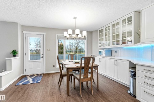 Open-concept dining and kitchen space featuring wood-finish flooring, white cabinetry with glass-front upper cabinets, light-toned countertops, and a full-light exterior door - 14755 131 Street Nw, Edmonton, AB - Indoor Photo Showing Dining Room