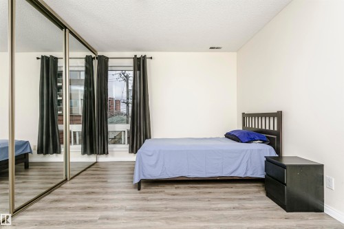 Room featuring light wood-finish flooring, a mirrored closet, white walls, and a window with dark curtains - 303 10026 112 Street, Edmonton, AB - Indoor Photo Showing Bedroom