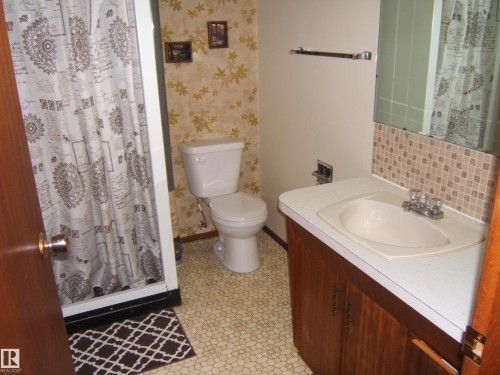 Bathroom featuring a white toilet, integrated sink with chrome faucet, and a mirror with tile backsplash - 5030 52 Avenue, Andrew, AB - Indoor Photo Showing Bathroom