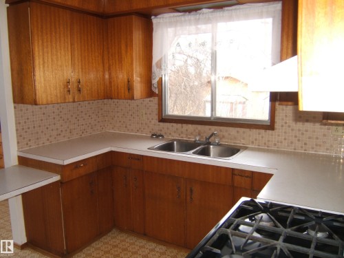 Kitchen featuring wood-finish cabinetry, a double basin stainless steel sink, and a tile backsplash - 5030 52 Avenue, Andrew, AB - Indoor Photo Showing Kitchen With Double Sink