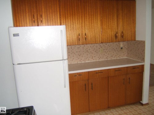 Kitchen area featuring wood-finish cabinetry, a white refrigerator, light-toned countertops, and a patterned tile backsplash - 5030 52 Avenue, Andrew, AB - Indoor Photo Showing Kitchen