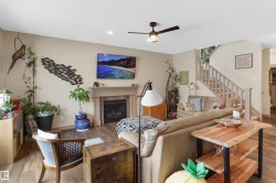 Living room featuring wood-finish flooring, a fireplace with a mantel, a ceiling fan, and a light-toned staircase with wood railings - 