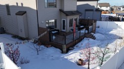 Rear exterior featuring a multi-level wood-finish deck with glass panel railings, direct access from the interior, and stairs descending to the yard - 