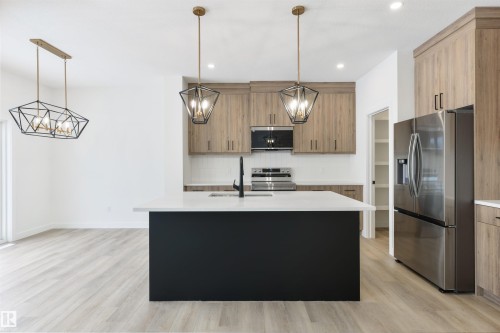 Open-concept kitchen featuring a black island with a white countertop, stainless steel appliances, wood-finish cabinetry, and contemporary pendant lighting - 162 Caldeon Crescent, Spruce Grove, AB - Indoor Photo Showing Kitchen With Upgraded Kitchen