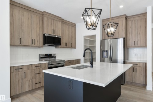 Kitchen featuring a central island with a dual-basin sink and matte black faucet, stainless steel appliances, wood-finish cabinetry with black hardware, white subway tile backsplash, and two geometric pendant lights - 162 Caldeon Crescent, Spruce Grove, AB - Indoor Photo Showing Kitchen With Double Sink With Upgraded Kitchen