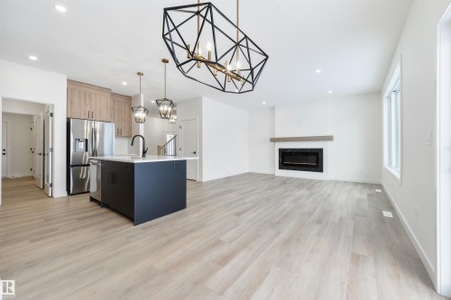 Open-concept living space featuring light wood-finish flooring, a modern electric fireplace with a wood mantle, a kitchen island with a dark base and light countertop, and stainless steel appliances - 162 Caldeon Crescent, Spruce Grove, AB - Indoor Photo Showing Kitchen With Upgraded Kitchen