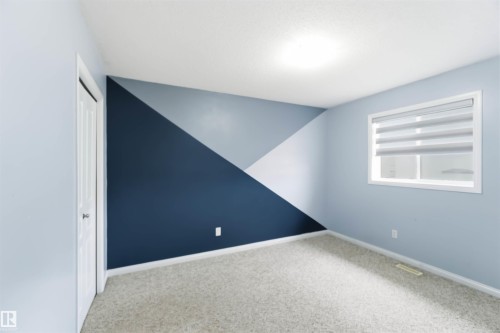 Interior room featuring a geometric accent wall in varying blue tones, light-colored carpet flooring, a white bi-fold closet door, and a window with horizontal blinds - 16412 60 Street, Edmonton, AB - Indoor Photo Showing Other Room