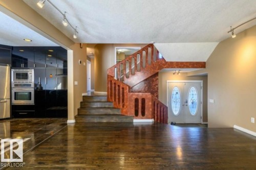 Entryway featuring a double-door entrance with oval glass inserts, a prominent staircase with decorative railing, wood-finish flooring, track lighting, and a partial view of a kitchen with dark cabinetry and stainless steel appliances - 18633 122 Avenue, Edmonton, AB - Indoor Photo Showing Other Room