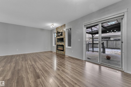 Spacious room featuring wood-finish flooring, a stone accent wall with a built-in electric fireplace, and a large sliding glass door opening to a private wooden deck with a covered pergola - 718 177 Street, Edmonton, AB - Indoor With Fireplace