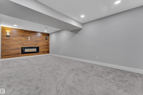 Spacious room featuring a wood-finish accent wall with an inset electric fireplace, two wall-mounted sconces, recessed lighting, and light gray carpeting - 718 177 Street, Edmonton, AB - Indoor