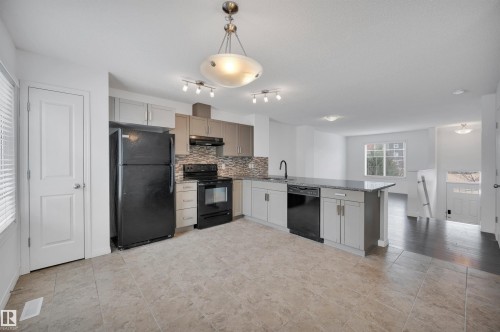 Open-concept kitchen featuring an island with a breakfast bar, stone countertops, stainless steel sink with a gooseneck faucet, black appliances, and tile flooring - 13 1030 Chappelle Boulevard, Edmonton, AB - Indoor Photo Showing Kitchen