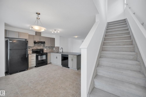 Open-concept kitchen featuring gray cabinetry, a mosaic tile backsplash, black appliances, a central island with a sink, and tile flooring - 13 1030 Chappelle Boulevard, Edmonton, AB - Indoor Photo Showing Kitchen