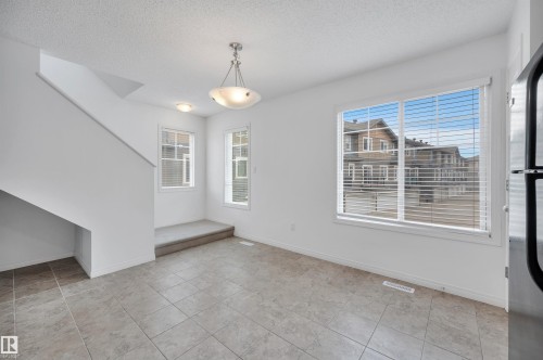 Bright open-concept interior featuring large windows, neutral-toned tile flooring, white walls, and a contemporary chandelier - 13 1030 Chappelle Boulevard, Edmonton, AB - Indoor Photo Showing Other Room