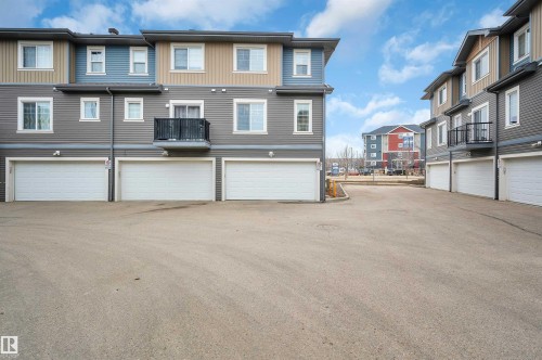 Multi-level townhome exterior featuring horizontal siding in shades of grey, blue, and tan, individual white garage doors, and a private balcony with black metal railings - 13 1030 Chappelle Boulevard, Edmonton, AB - Outdoor With Facade