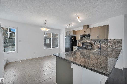 Kitchen featuring a granite-topped island, black appliances, textured tile backsplash, and light-toned cabinetry - 13 1030 Chappelle Boulevard, Edmonton, AB - Indoor Photo Showing Kitchen With Double Sink