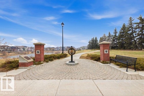 Paved stone pathway leading to a landscaped park area with a water feature, complete with a street lamp, brick columns, and a park bench - 18, 2560 Pegasus Boulevard, Edmonton, AB - Outdoor