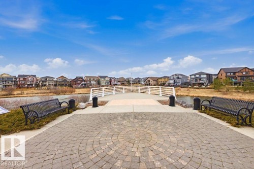 Paved walkway featuring a circular paver design, two black metal benches, and a white pedestrian bridge over a body of water - 18, 2560 Pegasus Boulevard, Edmonton, AB - Outdoor With View