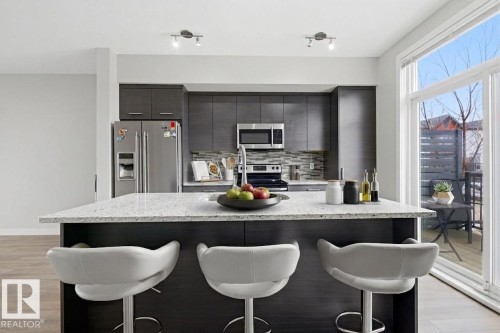 Modern kitchen featuring an island with a light-toned countertop, dark wood-finish cabinetry, stainless steel appliances, a mosaic tile backsplash, and wood-finish flooring - 18, 2560 Pegasus Boulevard, Edmonton, AB - Indoor Photo Showing Kitchen With Upgraded Kitchen