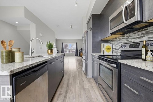 Contemporary kitchen featuring granite-look countertops, stainless steel appliances, a mosaic tile backsplash, wood-finish flooring, and dark cabinetry - 18, 2560 Pegasus Boulevard, Edmonton, AB - Indoor Photo Showing Kitchen With Double Sink With Upgraded Kitchen