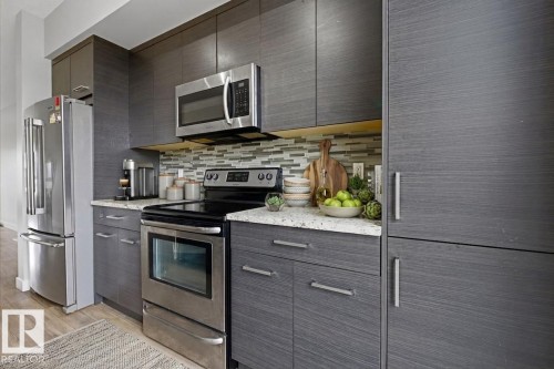 Contemporary kitchen featuring stainless steel appliances, dark wood-finish cabinetry, light speckled countertops, and a mosaic tile backsplash - 18, 2560 Pegasus Boulevard, Edmonton, AB - Indoor Photo Showing Kitchen