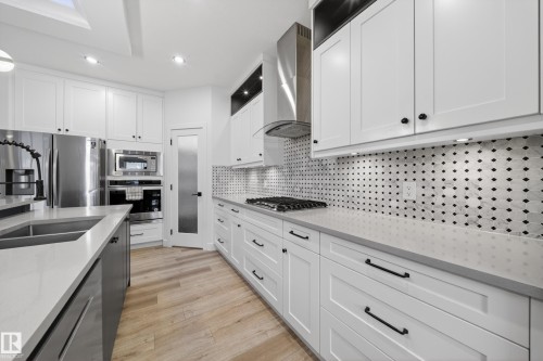 Modern kitchen featuring white shaker cabinetry, stainless steel appliances, a geometric tile backsplash, light-toned wood-finish flooring, and a large island with an undermount sink - 7414 Chivers Crescent Sw, Edmonton, AB - Indoor Photo Showing Kitchen With Double Sink With Upgraded Kitchen