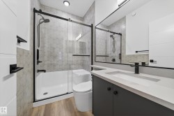 Bathroom featuring wood-finish flooring, a white vanity with an integrated sink and matte black faucet, a built-in bathtub with a clear glass sliding door, and patterned tile shower surround - 