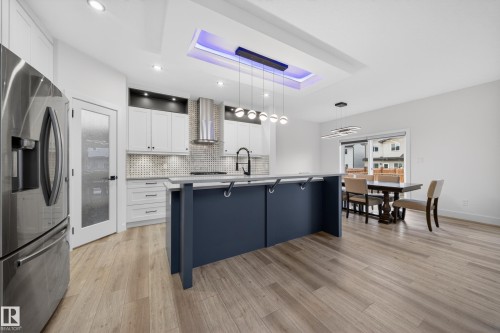 Open-concept kitchen and dining area featuring light wood-finish flooring, a kitchen island with a built-in sink, white cabinetry with a patterned backsplash, stainless steel appliances, and a tray ceiling with recessed lighting - 7414 Chivers Crescent Sw, Edmonton, AB - Indoor Photo Showing Kitchen With Upgraded Kitchen